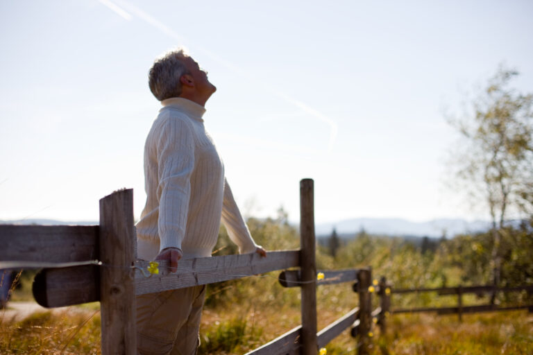 Älterer Mann lehnt an einem Holzzaun in einer sonnigen Naturlandschaft und atmet bewusst die frische Luft ein – Biohacking durch Natur und Achtsamkeit.